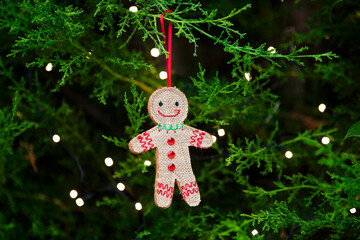 Gingerbread man handmade Christmas decoration hanging on branch or green tree with golden twinkling fairy lights 