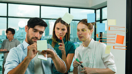 Professional business team brainstorm, sharing, thinking idea while skilled male leader write on sticky notes at glass wall. Skilled manager placed business plan on glass wall at office. Tracery