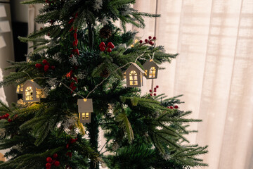 Close-up of a Christmas tree adorned with lit-up house ornaments, pine cones, and red berries, set against a softly lit curtain background