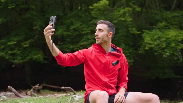 Fitness coach live streaming on phone in nature. Young man talking on a video call in a forest. Positive influencer creating content for social media outdoors.