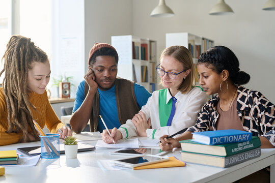 Young female teacher sitting at table with multiethnic group of students during class in school or college