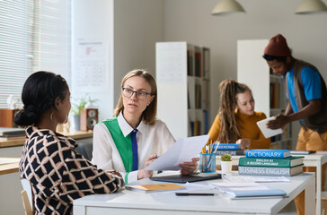 Young woman as female teacher talking with student while sitting at table during class in school or college