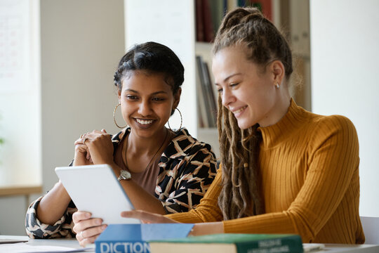 Two smiling young female students sitting at table and watching video on digital tablet while doing homework together in library