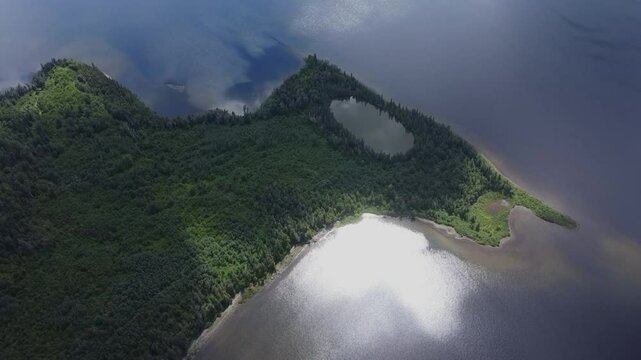 Un drone en haute altitude d&eacute;voile un petit lac ovale cach&eacute; dans la for&ecirc;t verdoyante refl&eacute;tant la lueur du soleil et les nuages en bordure d'un lac d'eau bleu. ( Seigneurie Lac Matap&eacute;dia, Qu&eacute;bec. )