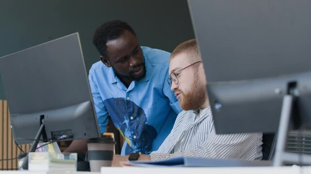 Two young ethnically diverse programmers having conversation while solving problem with code in office