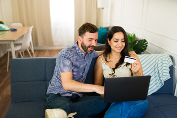 Young couple is sitting on a sofa at home, smiling while using a laptop and a credit card to shop online