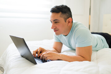 Profile view of a good looking Latin man lounging on his bed while using his laptop at home