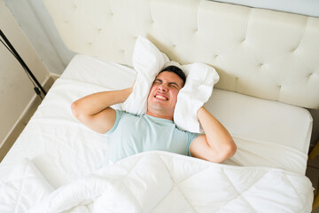 Stressed and upset man lying in bed and using a pillow to block out noisy sounds from his neighbors