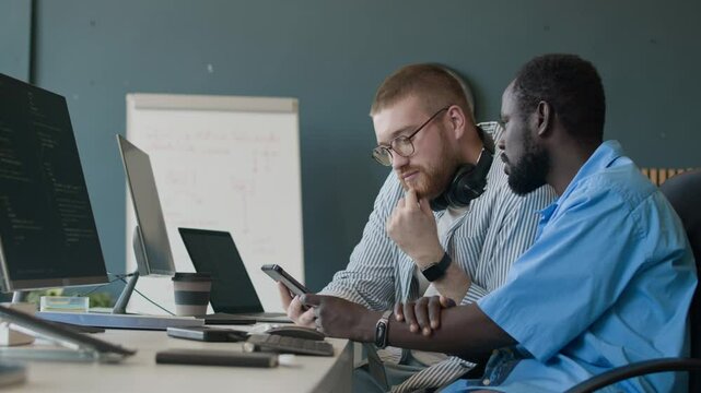 Side view shot of two gen Z ethnically diverse IT specialists working together on code in office, using digital tablet and computers