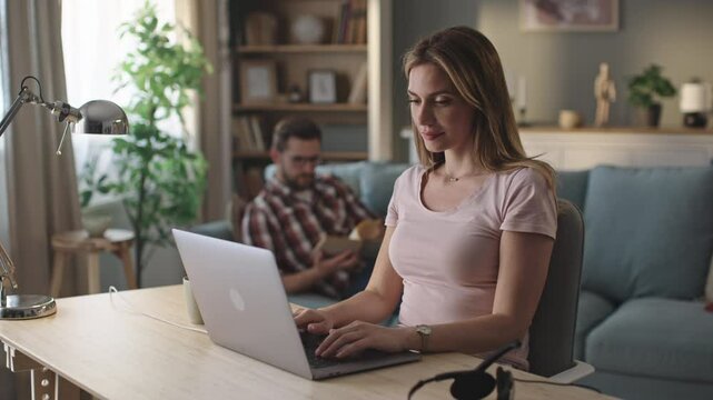 Business woman working online at home on weekend in slow-motion. Her husband lying on sofa and reading book in the background