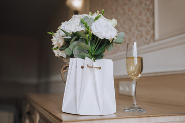 Gift - paper basket with fresh flowers and glass with champagne stands on wooden table indoors. Close-up photo, festive gift for birthday, wedding.