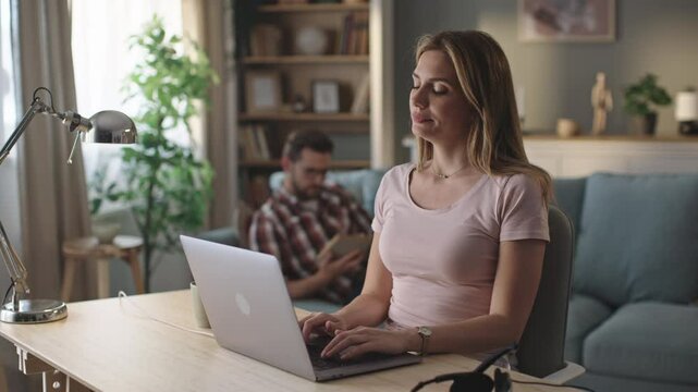 Beautiful tired female sitting at a desk in a cozy living room and using laptop at home. She is suffering from neck pain and massaging her neck