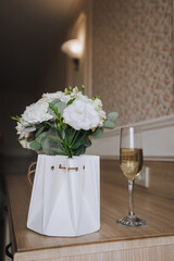 Gift - paper basket with fresh flowers and glass of champagne stands on wooden table indoors. Photo, holiday gift for birthday, wedding.
