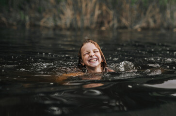 Happy beautiful girl, smiling child swims laughing in water on lake, river, sea in summer. Photography, close-up portrait, childhood concept.