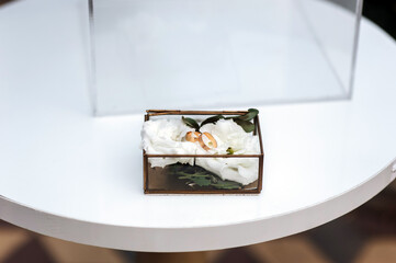 Beautiful glass box with golden wedding rings and white rose petals stands on wooden table at ceremony. Wedding photography, accessories, details.