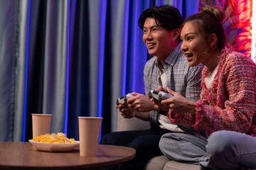 Sitting sofa of happy smiling couple playing video game control by joystick fronted snack and drinks competing challenge level with funny at modern neon blub light comfy living room at home. Infobahn.