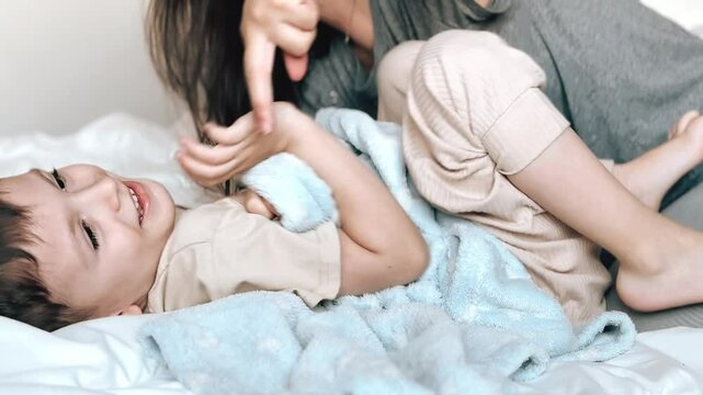 Mom tickles the feet of her little son, wrapped in a blue blanket. Mom and child have fun together in a cozy bedroom at home. Happy children with mother. Bright photos of the baby's childhood.
