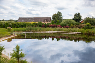 Obraz premium Peaceful rural landscape with a pond reflecting a barn and lush greenery in the Latvian countryside on a cloudy day.
