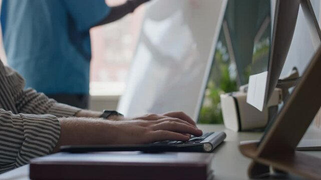 Hands of unrecognizable male programmer typing on desktop computer while his colleague writing on whiteboard in background