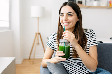 Happy young woman drinking healthy green juice sitting on sofa at home. Healthy lifestyle and diet...
