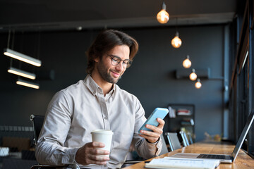 Smiling young adult European business man executive, busy businessman entrepreneur sitting at desk in office holding smartphone using mobile cell phone managing digital apps on cellphone at work.