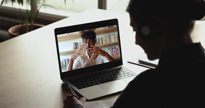 Indian schoolmates studying together using video call app, laptop screen view over female student shoulder writing in copybook, make notes, e-learning with help of on-line teacher. Education, tech