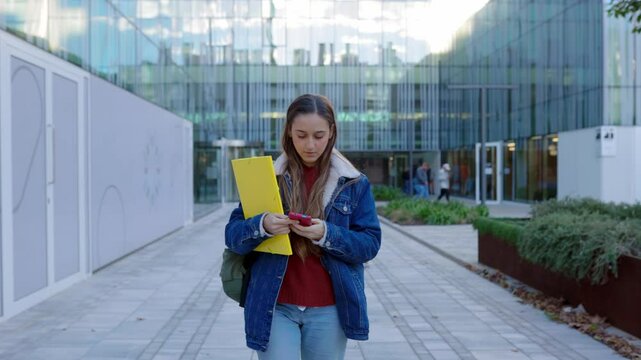 Young caucasian student girl using mobile phone while leaving university campus after classes. Education and technology concept.