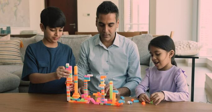 Focused Indian dad and two kids constructing city from toy blocks, building towers in home living room, talking to preteen son and little daughter girl, smiling, laughing