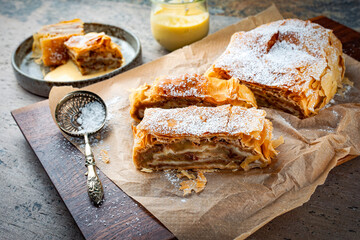 Traditional Bavarian apfelstrudel with backed apples, raisins and icing sugar served as close-up on a wooden design board with backing paper