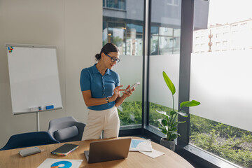 A professional woman is engaged with her smartphone while working in a modern office filled with greenery