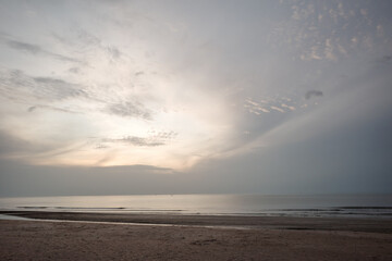 A tranquil beach scene captured during sunset, featuring a vast, soft-toned sky with light clouds and a calm, reflective sea. 