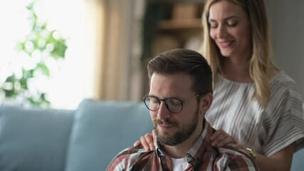 Caucasian man leaning back on his girlfriend sitting on couch in living room behind him, massaging neck and talking happily. Shot in slow-motion