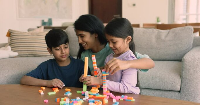 Happy young Indian mom helping cute sibling kids to stack towers from toy colorful bricks, playing learning creative games with son and little daughter in home living room. Nanny watching girl and boy