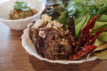 A close-up shot of a delicious Thai spicy sausage garnished with crispy fried garlic and herbs, served alongside fresh green leaves and dried red chilies. 