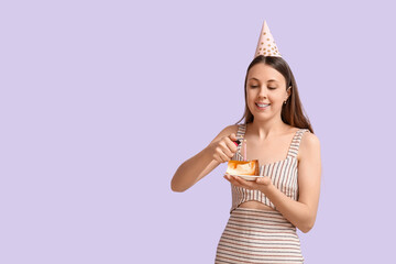 Beautiful young woman lighting candle on tasty birthday cake against lilac background