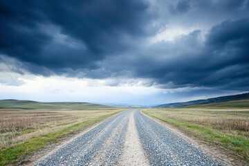 Fototapeta premium Low angle view of gravel road through countryside meadows, captured under a cloudy sky at spring evening.