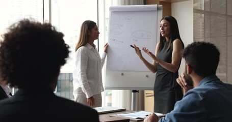 Two positive successful business women presenting project to team of colleagues, speaking at flipchart in boardroom, pointing at white board, smiling, coaching employees, offering work strategy - Powered by Adobe