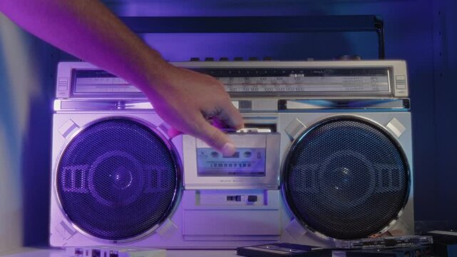 A hand carefully places a cassette tape into a vintage music player and turns it on in a studio bathed in neon light. The close-up shot captures the essence of retro audio technology.