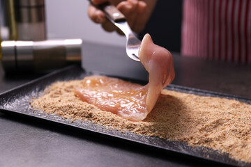 Making schnitzel. Woman coating slice of meat with bread crumbs at dark table, closeup