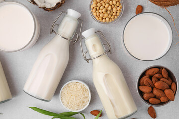 Different types of vegan milk with ingredients and leaves on light grey table, flat lay