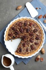 Delicious pecan pie in baking dish, caramel sauce, cake server and fresh nuts on gray textured table, flat lay