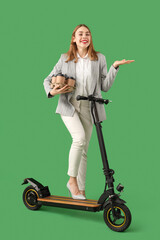 Portrait of young businesswoman with takeaway coffee cups and kick scooter showing something on green background © Pixel-Shot