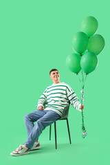 Young man with bunch of balloons sitting on chair against green background