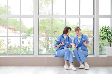 Female medical interns with clipboards sitting on windowsill in clinic