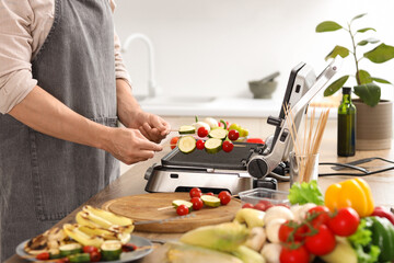 Young man cooking tasty vegetables skewers on modern electric grill in kitchen, closeup