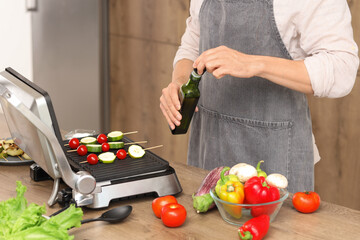 Young man adding olive oil onto tasty vegetables skewers on modern electric grill in kitchen,...