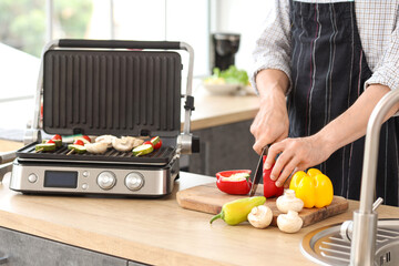 Young man cutting fresh vegetables to cooking on modern electric grill in kitchen, closeup
