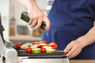 Young man adding olive oil onto tasty vegetables skewers on modern electric grill in kitchen,...