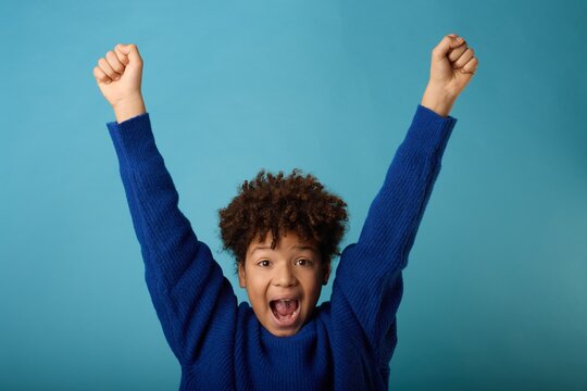 A joyful young african american boy celebrating with hands raised against a bright blue background