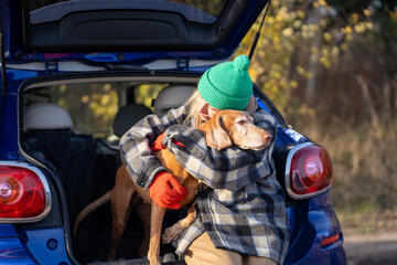 Loving woman dog owner snuggling hugging pet after autumn forest walk resting together. Close friendship bond between caring female and canine vizsla, sitting with embrace leaning on trunk of car. 
